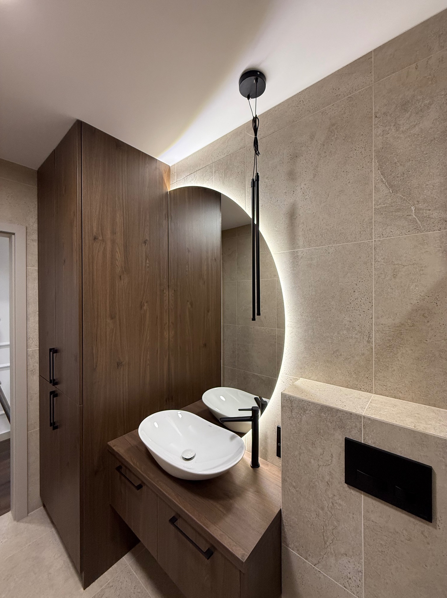 Nowoczesna łazienka z umywalką nablatową, okrągłym lustrem z podświetleniem LED i szafką z ciemnego drewna. Beżowe płytki na ścianach i podłodze.