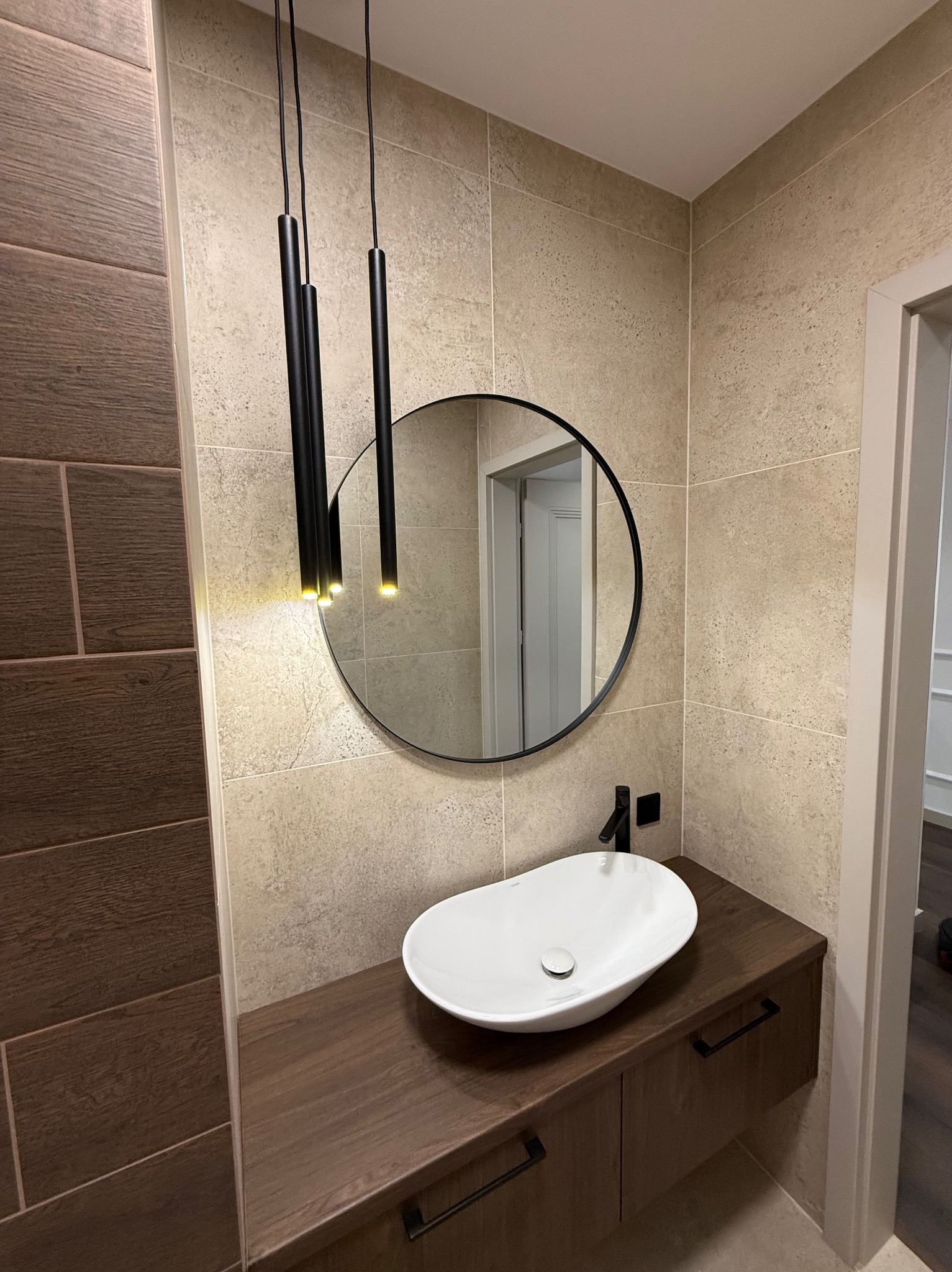 Nowoczesna łazienka z owalną umywalką, okrągłym lustrem i wiszącymi lampami. Beżowe płytki na ścianach i drewnopodobna szafka pod umywalką tworzą spójny design.