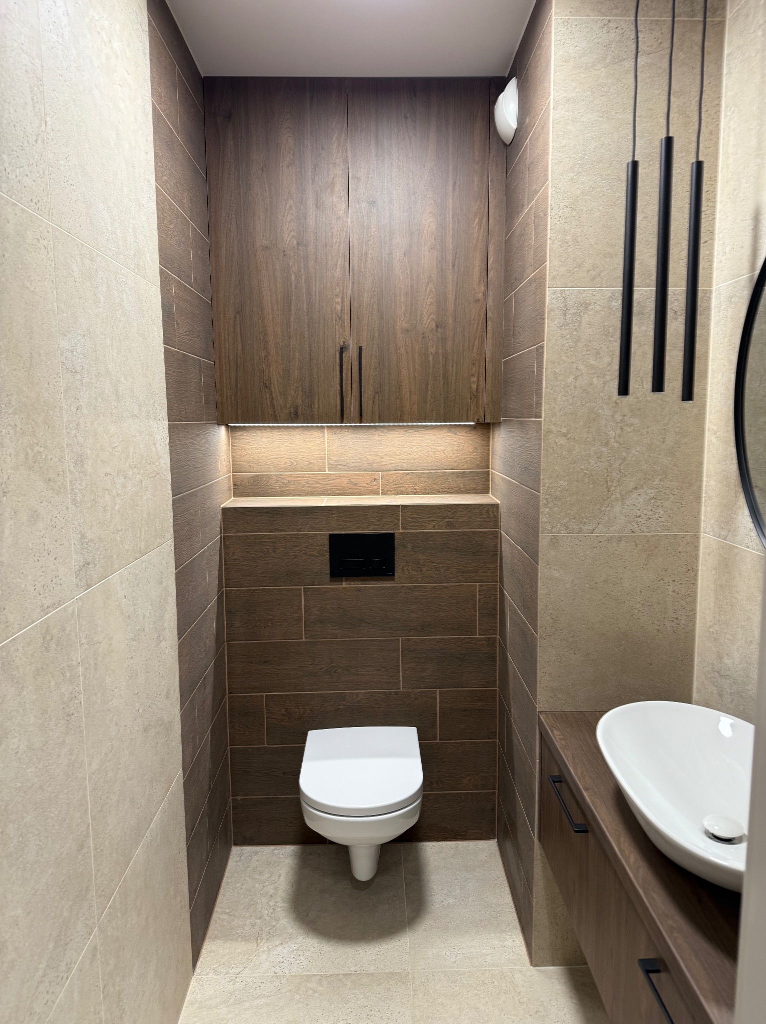 Nowoczesna łazienka po remoncie: beżowe i brązowe płytki, podwieszana toaleta, szafka z umywalką i wiszące lampy. Minimalistyczny design z akcentami drewna.