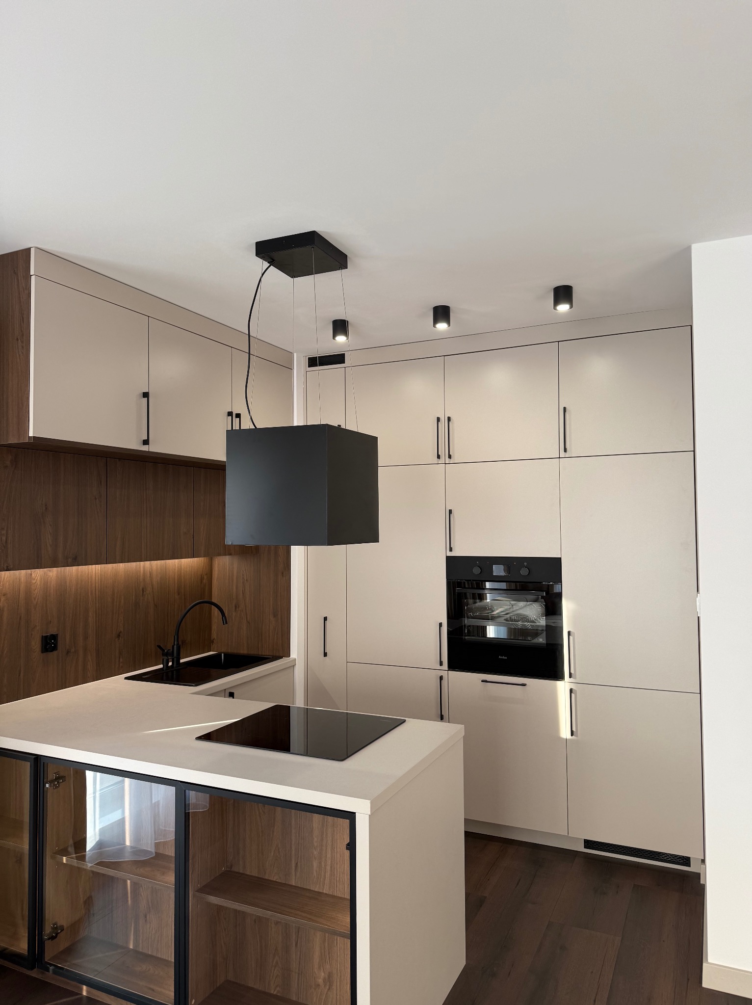 Nowoczesna kuchnia z wyspą, fronty szafek w jasnym odcieniu, czarne akcenty: okap, bateria, płyta indukcyjna. Drewniane wykończenie ściany nad blatem. Minimalistyczne oświetlenie.
