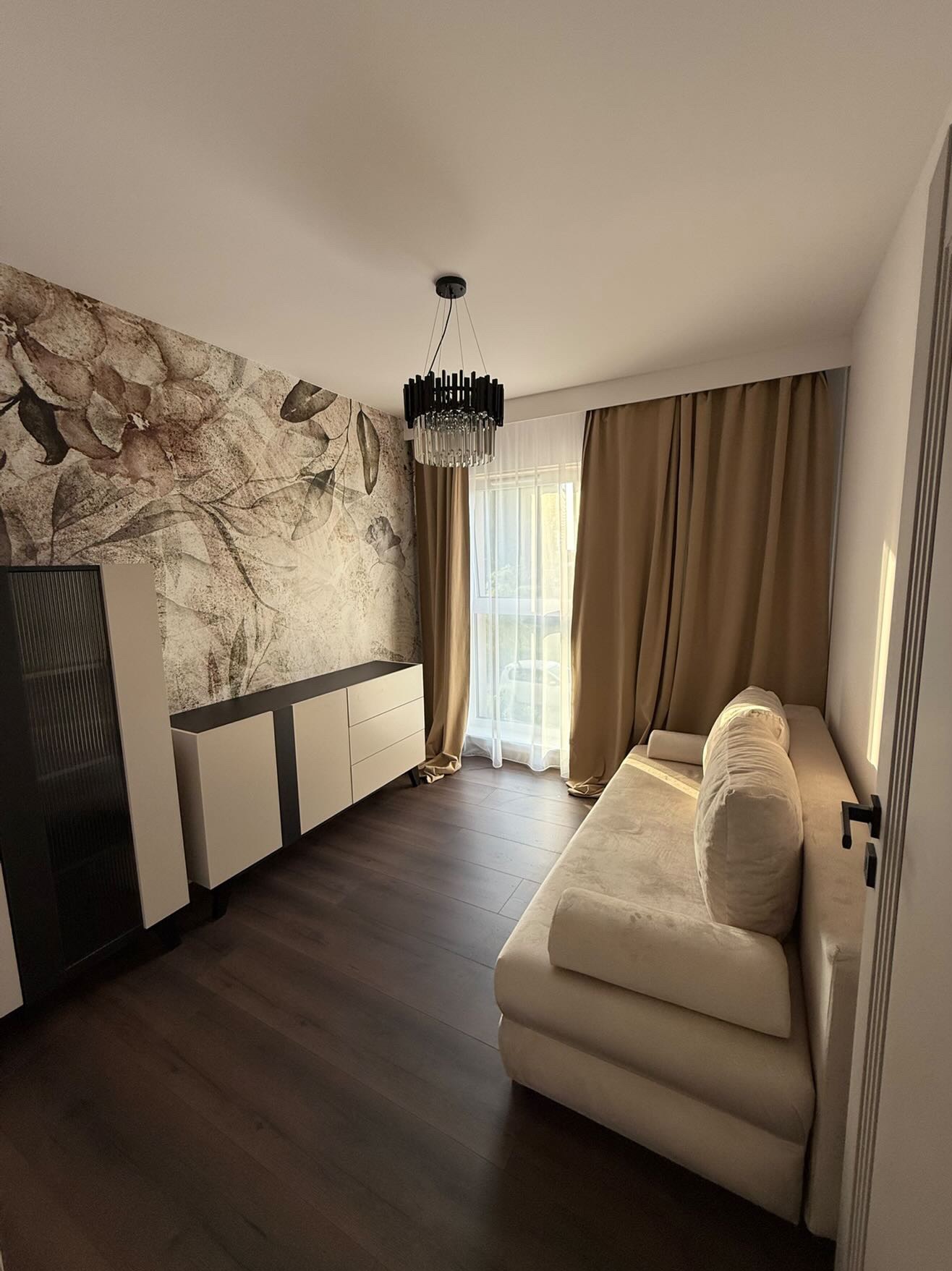 Elegancki salon z sofą, komodą i fototapetą w kwiaty. Ciemna podłoga i nowoczesny żyrandol. Stonowana kolorystyka wnętrza.