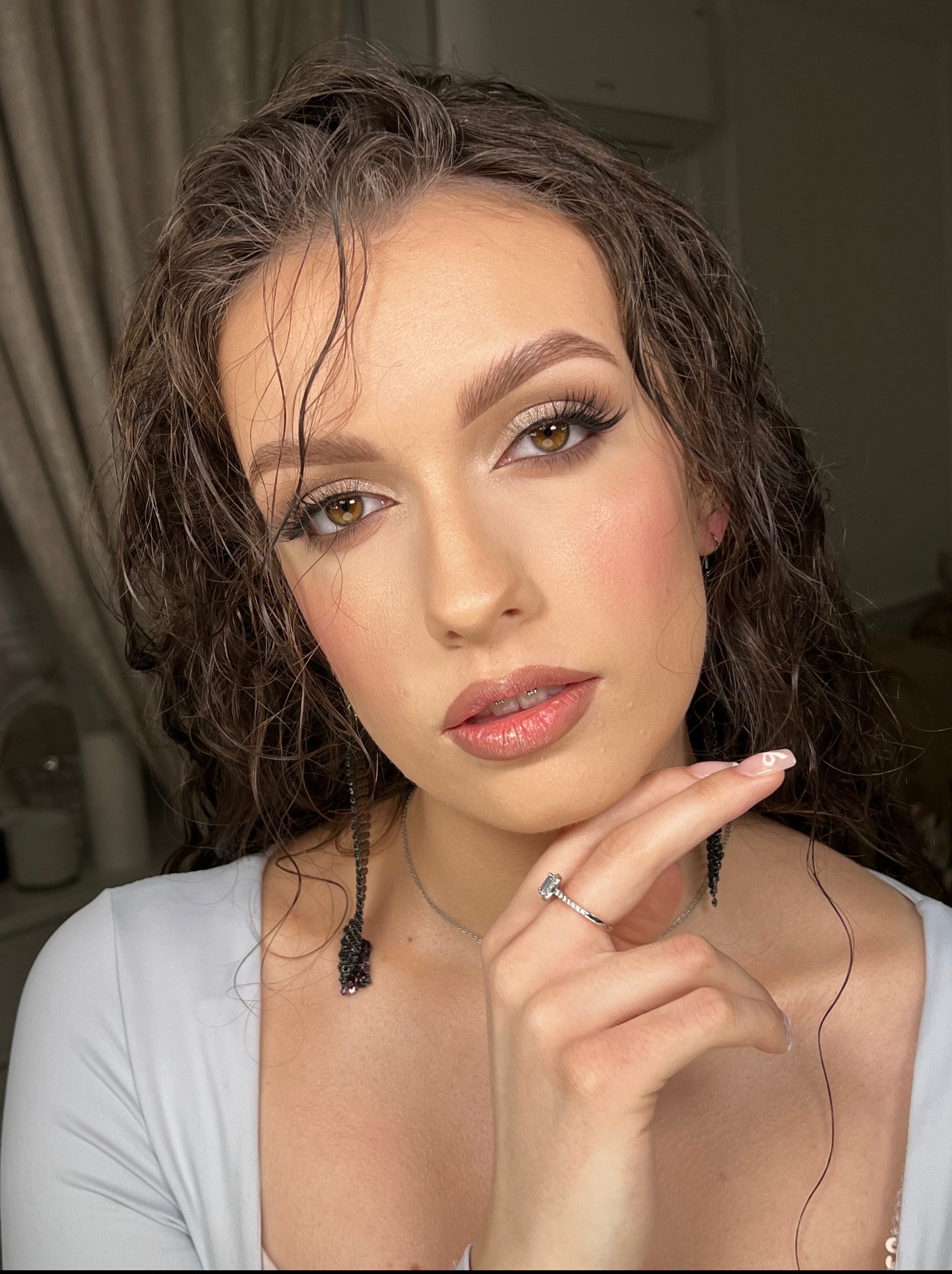 Portret kobiety z błyszczącym makijażem oczu i ust. Uwagę przykuwają długie, czarne kolczyki i pierścionek. Włosy lekko wilgotne.