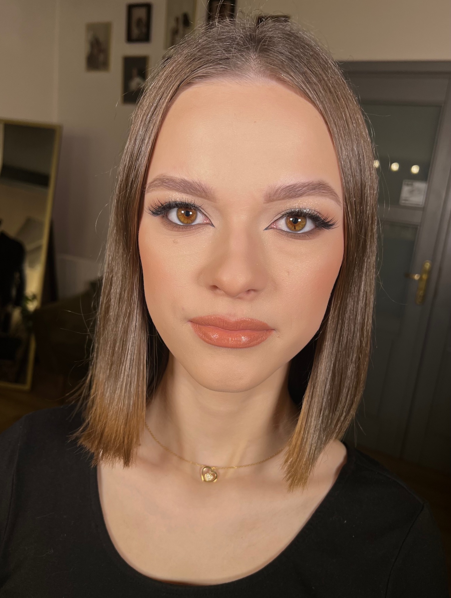 Portret kobiety z profesjonalnym makijażem: błyszczące cienie, sztuczne rzęsy, podkreślone brwi i usta w odcieniu nude. Złoty naszyjnik z sercem.
