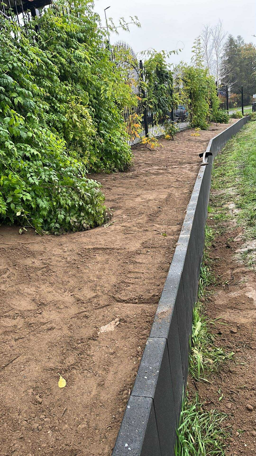 Ukończona ścieżka z obrzeżami z kostki, widok z góry, po prawej trawa, po lewej krzewy i czarne ogrodzenie. Ścieżka z ziemi.