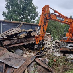 FHU SŁAWOMIR STARZYŃSKI - Pomarańczowa koparka Doosan na tle gruzów i zniszczonego budynku. Widoczne elementy konstrukcyjne i cegły. Prace rozbiórkowe w toku, pochmurne niebo.