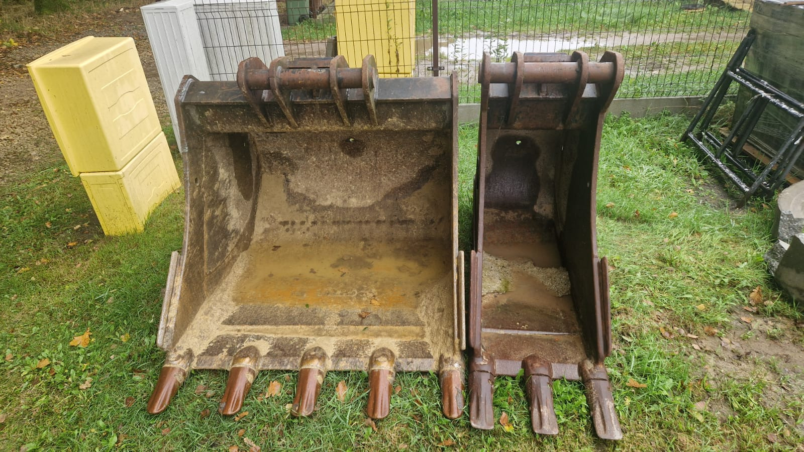 Dwie brudne łyżki do koparki, różnej wielkości, stoją na trawie. W tle widoczne elementy ogrodzenia i palety. Widok z bliska, z ostrym światłem.