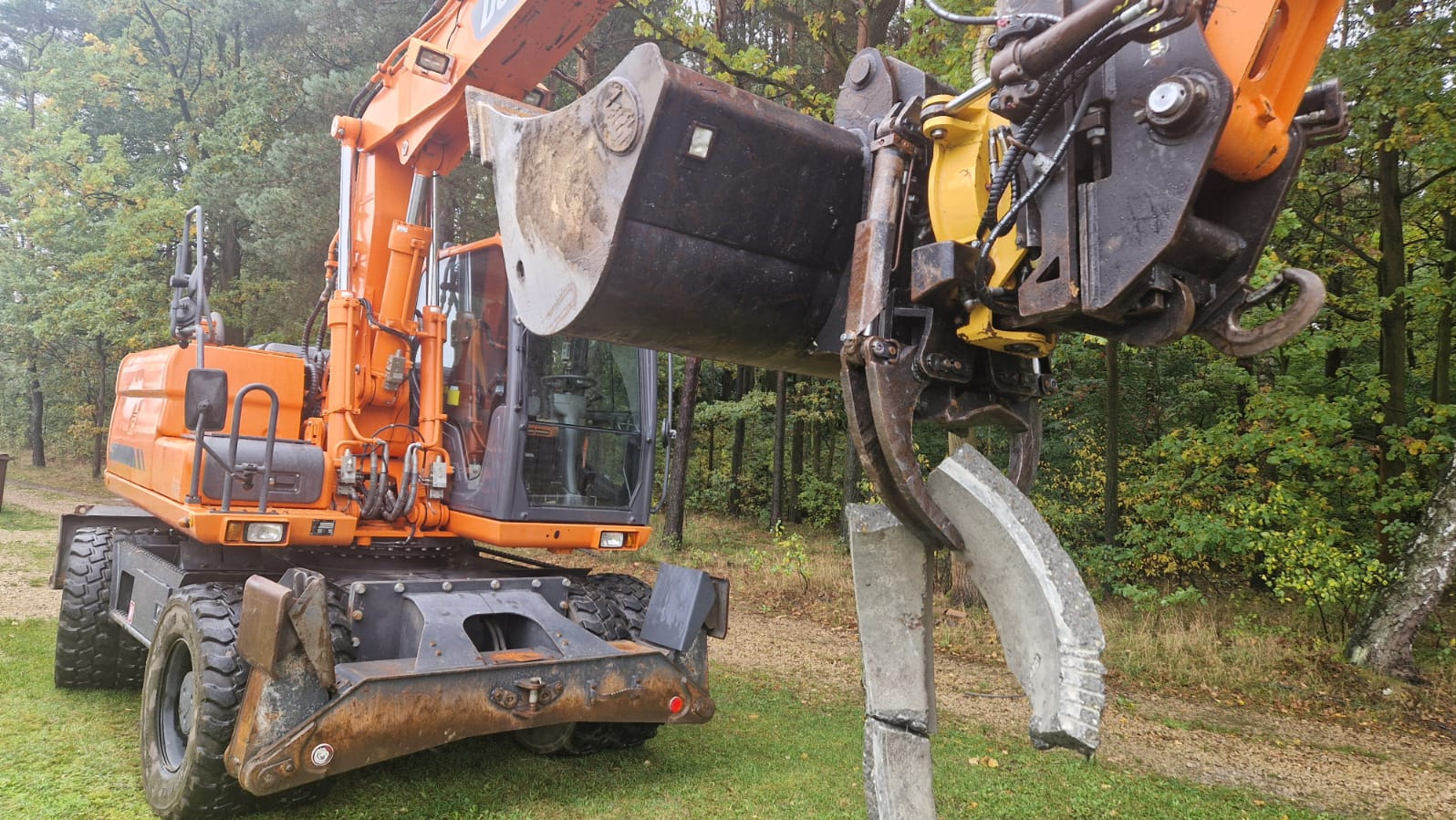 Pomarańczowa koparka kołowa z łyżką i chwytakiem podnosi fragment betonowego słupa na tle zieleni. Widoczne drzewa i trawa. Sprzęt ciężki w trakcie pracy.