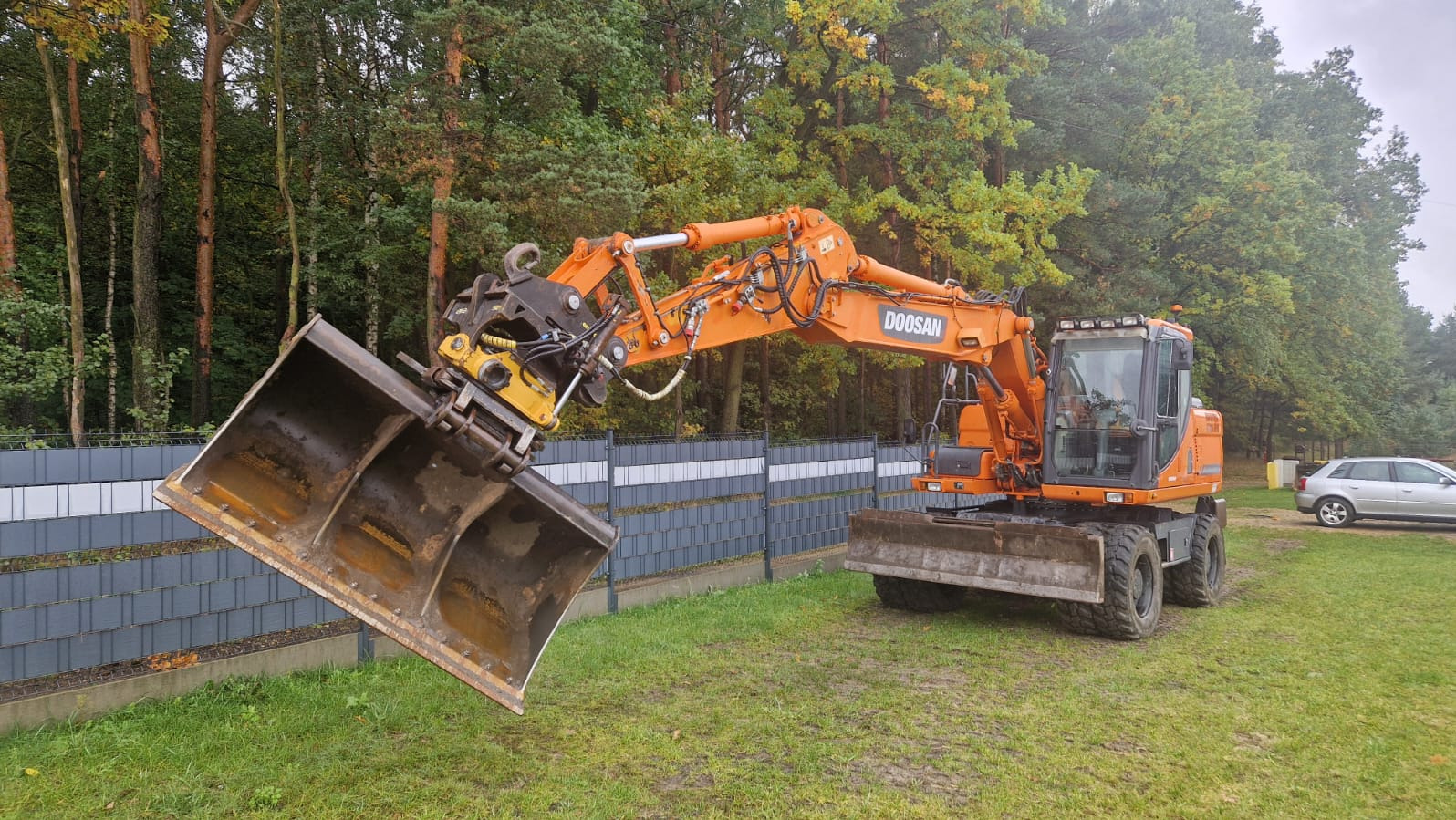 Pomarańczowa koparka Doosan z łyżką pochyloną na tle zieleni i szarego ogrodzenia. Na trawie ślady kół. W tle drzewa i zaparkowany samochód osobowy.