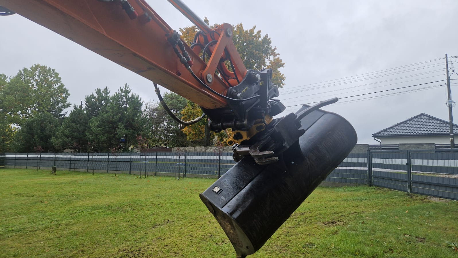 Pomarańczowe ramię koparki z czarną łyżką wbitą w zieloną trawę, na tle ogrodzenia i drzew. Widoczne ślady opon na trawie. Pochmurne niebo.