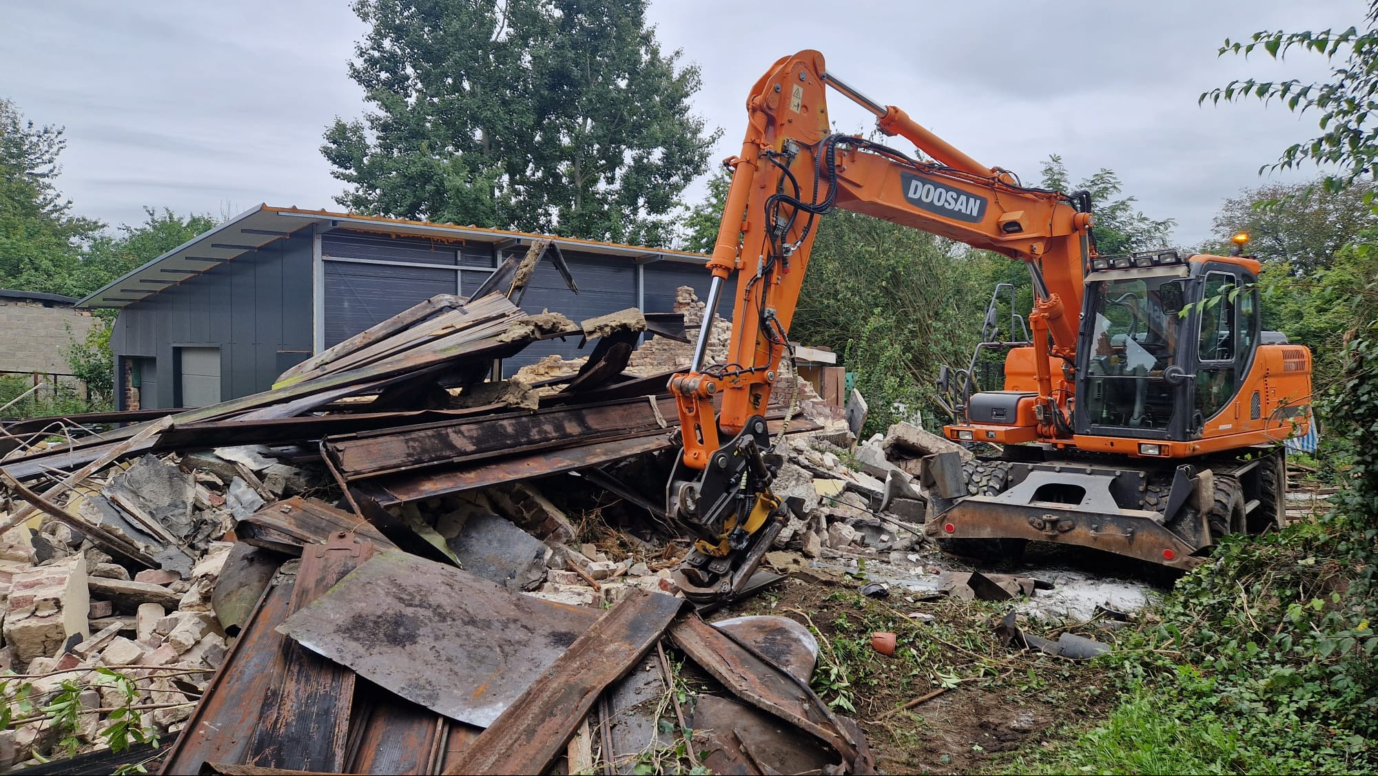 Pomarańczowa koparka Doosan na tle gruzów i zniszczonego budynku. Widoczne elementy konstrukcyjne i cegły. Prace rozbiórkowe w toku, pochmurne niebo.