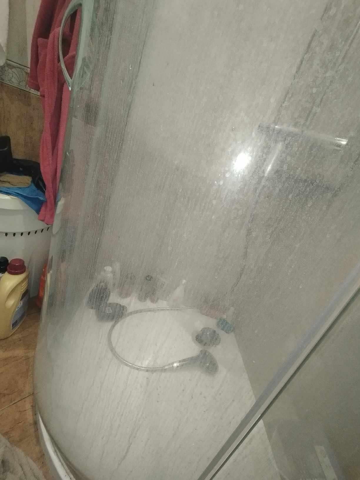 Zabrudzona kabina prysznicowa z widocznymi osadami i zaciekami na szklanych ścianach. Wewnątrz widoczne kosmetyki i słuchawka prysznicowa.