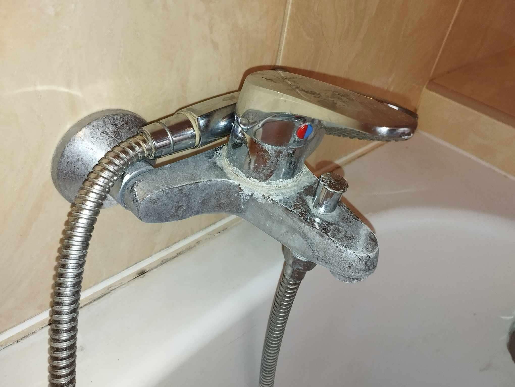 Zanieczyszczona kamieniem bateria wannowa z wężem prysznicowym na tle jasnych płytek. Widoczne osady wapienne na chromowanych elementach.