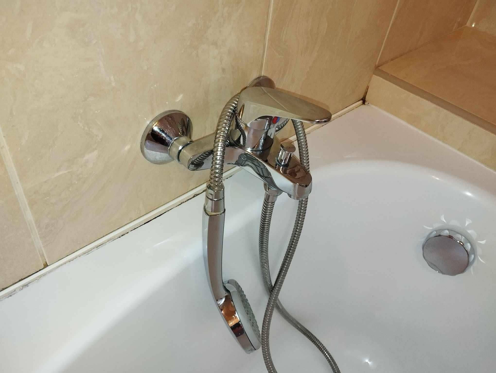 Bateria wannowa z chromowanym wykończeniem i słuchawką prysznicową na tle beżowych płytek i białej wanny. Widoczne ślady brudu i pleśni w fugach.