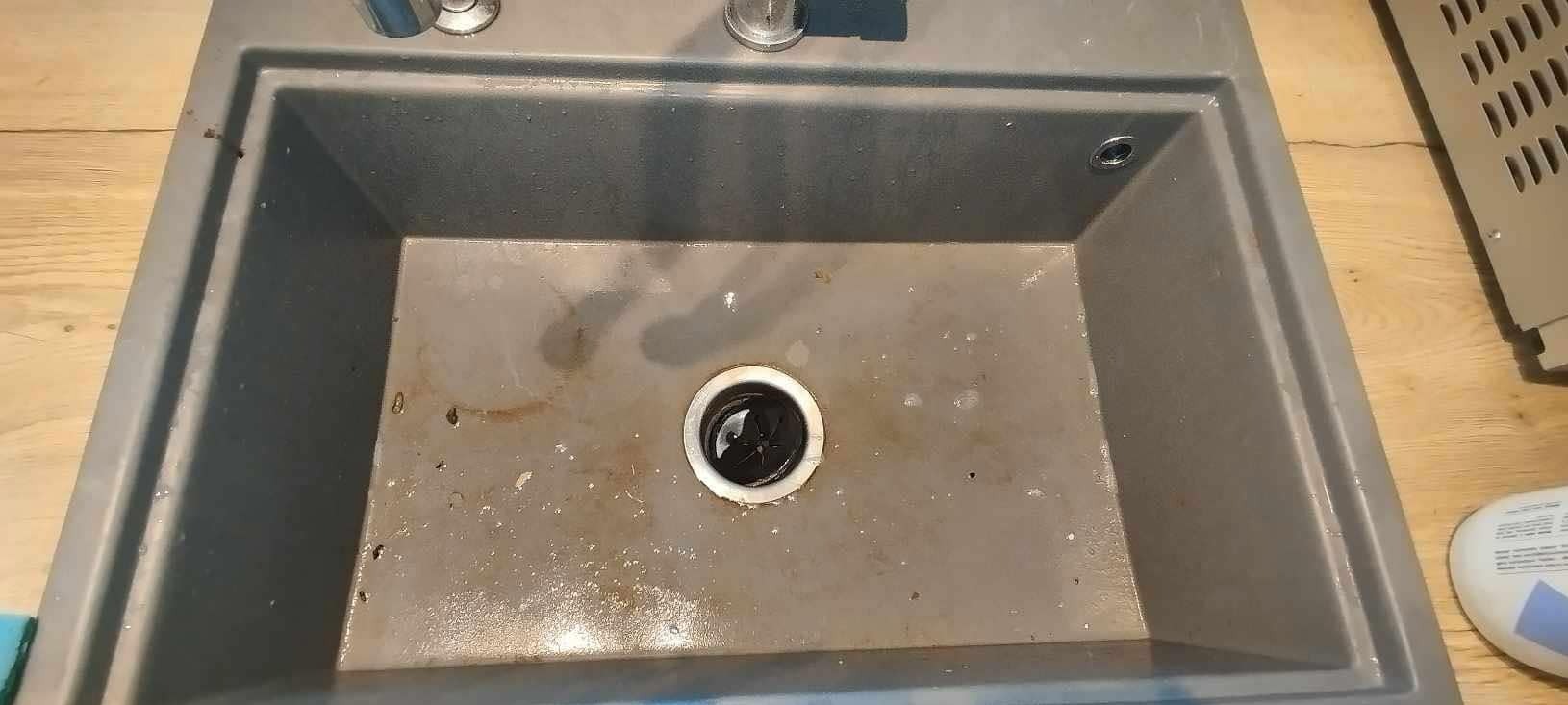 Szary, brudny zlew granitowy z widocznymi plamami i osadem, oczekujący na wyczyszczenie. Widoczny odpływ i fragment blatu. Ujęcie z góry.