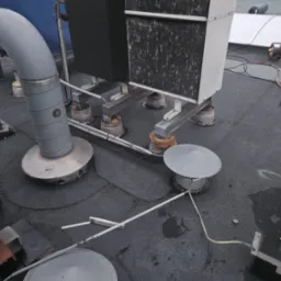 Instalacja na płaskim dachu z widocznymi elementami wentylacji i klimatyzacji. Pracownik z narzędziem w tle. Dach pokryty papą. Widoczne przewody i elementy mocujące.