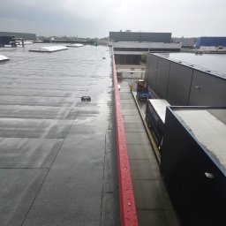 Michalina Frandzel - Widok z góry na płaski, mokry dach przemysłowy z odpływem i czerwoną barierką; w tle budynki i pochmurne niebo. Widoczny fragment dachu i rampy załadunkowej.