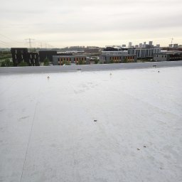 Michalina Frandzel - Biała, płaska powierzchnia dachu z widocznymi znakami ostrzegawczymi, budynkami w tle i pochmurnym niebem. Nowe krycie dachu wykonane z membrany PVC.