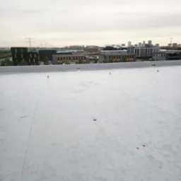 Biała, płaska powierzchnia dachu z widocznymi znakami ostrzegawczymi, budynkami w tle i pochmurnym niebem. Nowe krycie dachu wykonane z membrany PVC.