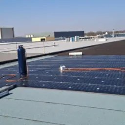 Prace dekarskie na dachu budynku: dekarz z palnikiem, butle gazowe, rolki papy. Widoczne warstwy izolacyjne i materiały przygotowane do montażu na jasnym dachu.