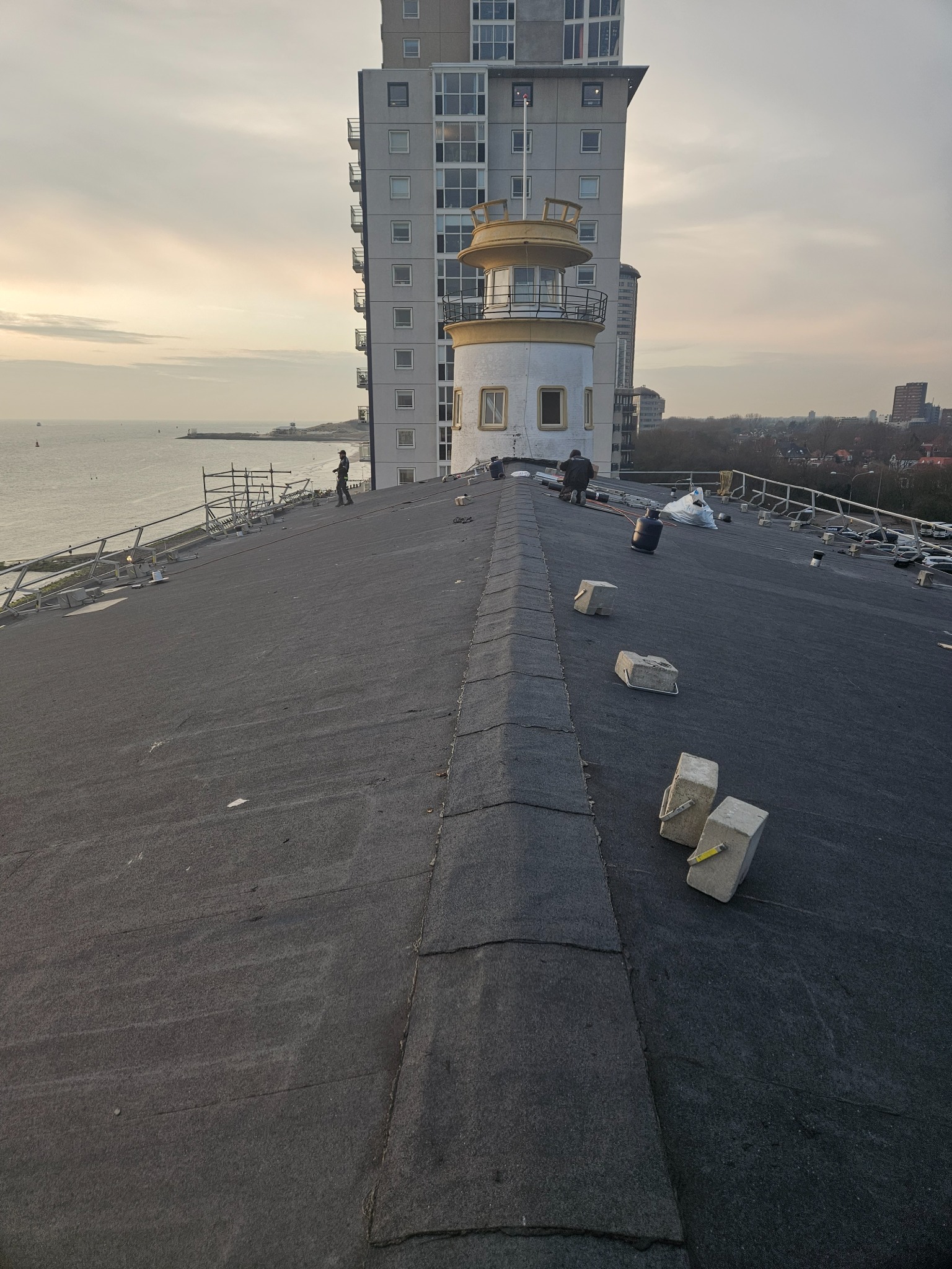 Praca na dachu budynku z wieżyczką widokową. Ekipa montuje papę na dużej powierzchni. W tle morze i budynki miejskie. Widoczne narzędzia i materiały budowlane.