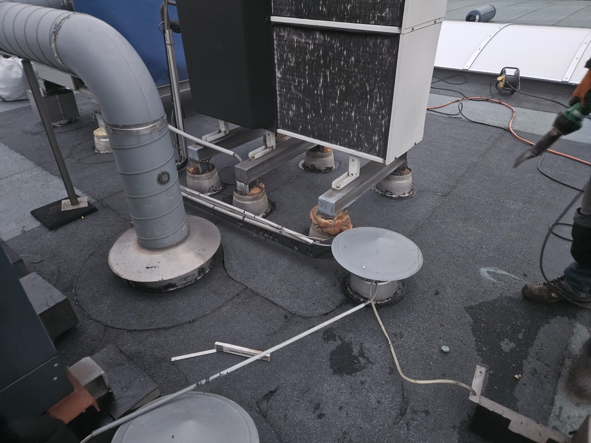 Instalacja na płaskim dachu z widocznymi elementami wentylacji i klimatyzacji. Pracownik z narzędziem w tle. Dach pokryty papą. Widoczne przewody i elementy mocujące.