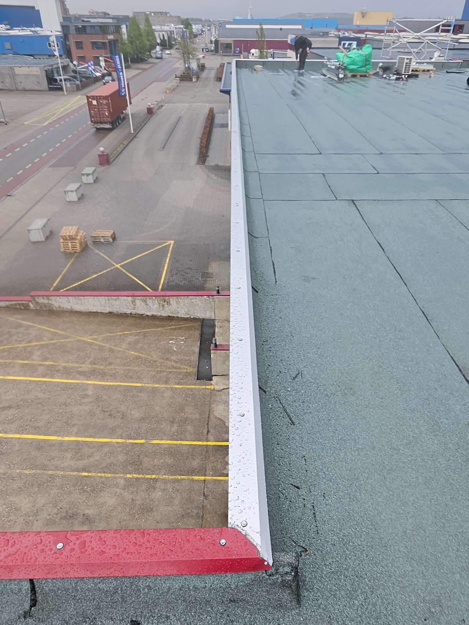 Widok z góry na fragment płaskiego dachu z papy termozgrzewalnej w trakcie prac. Widoczny fragment dachu, krawędź z obróbką blacharską i osoba pracująca na dachu.