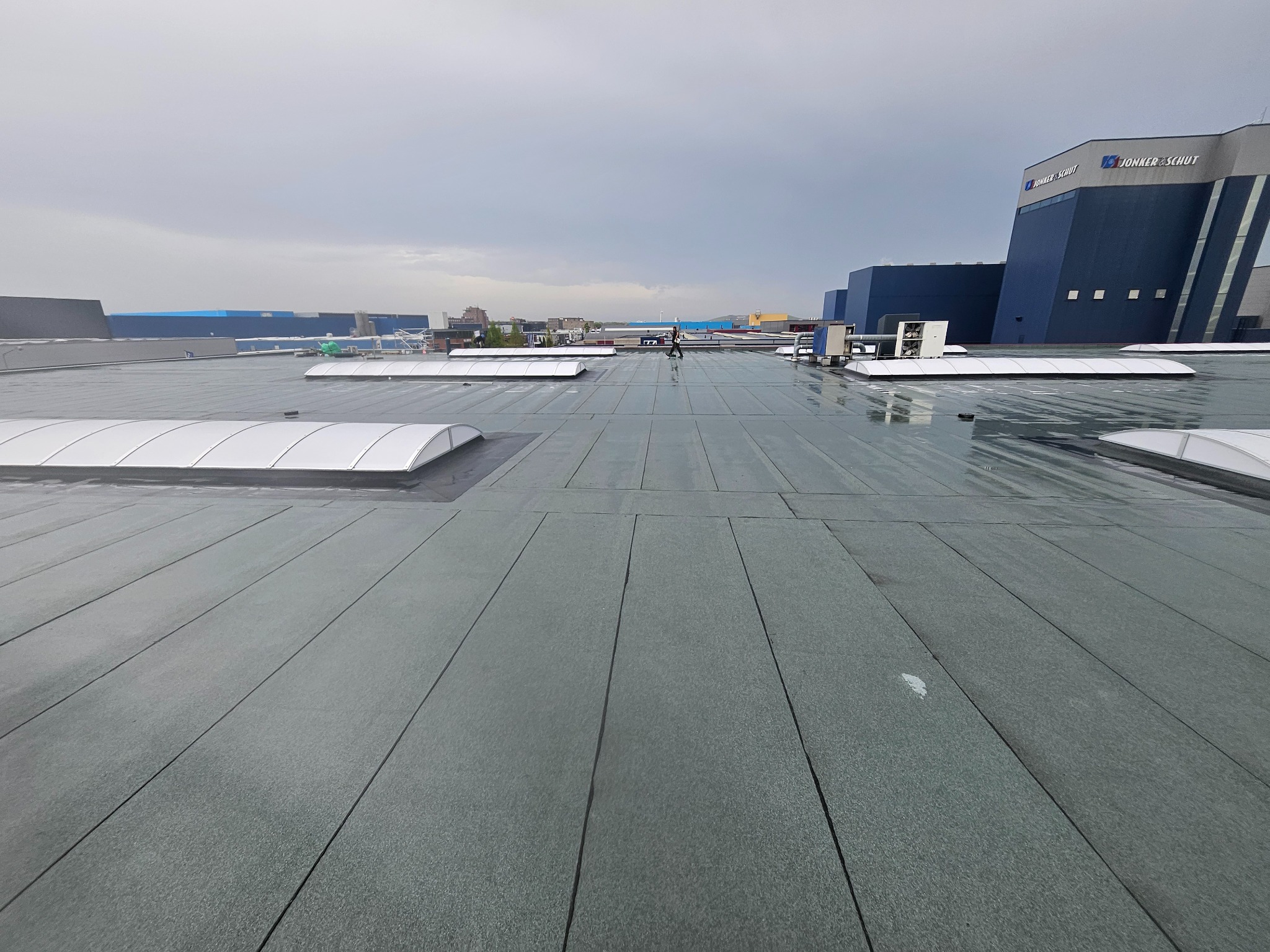 Płaski, pokryty papą dach budynku komercyjnego z świetlikami, widok z góry, w tle budynki przemysłowe pod pochmurnym niebem. Minimalistyczny design.