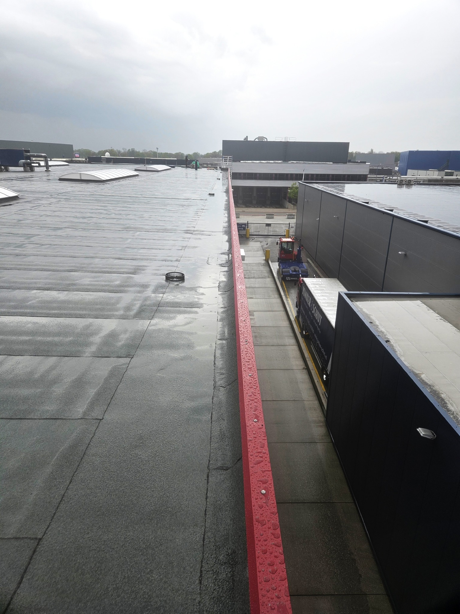 Widok z góry na płaski, mokry dach przemysłowy z odpływem i czerwoną barierką; w tle budynki i pochmurne niebo. Widoczny fragment dachu i rampy załadunkowej.