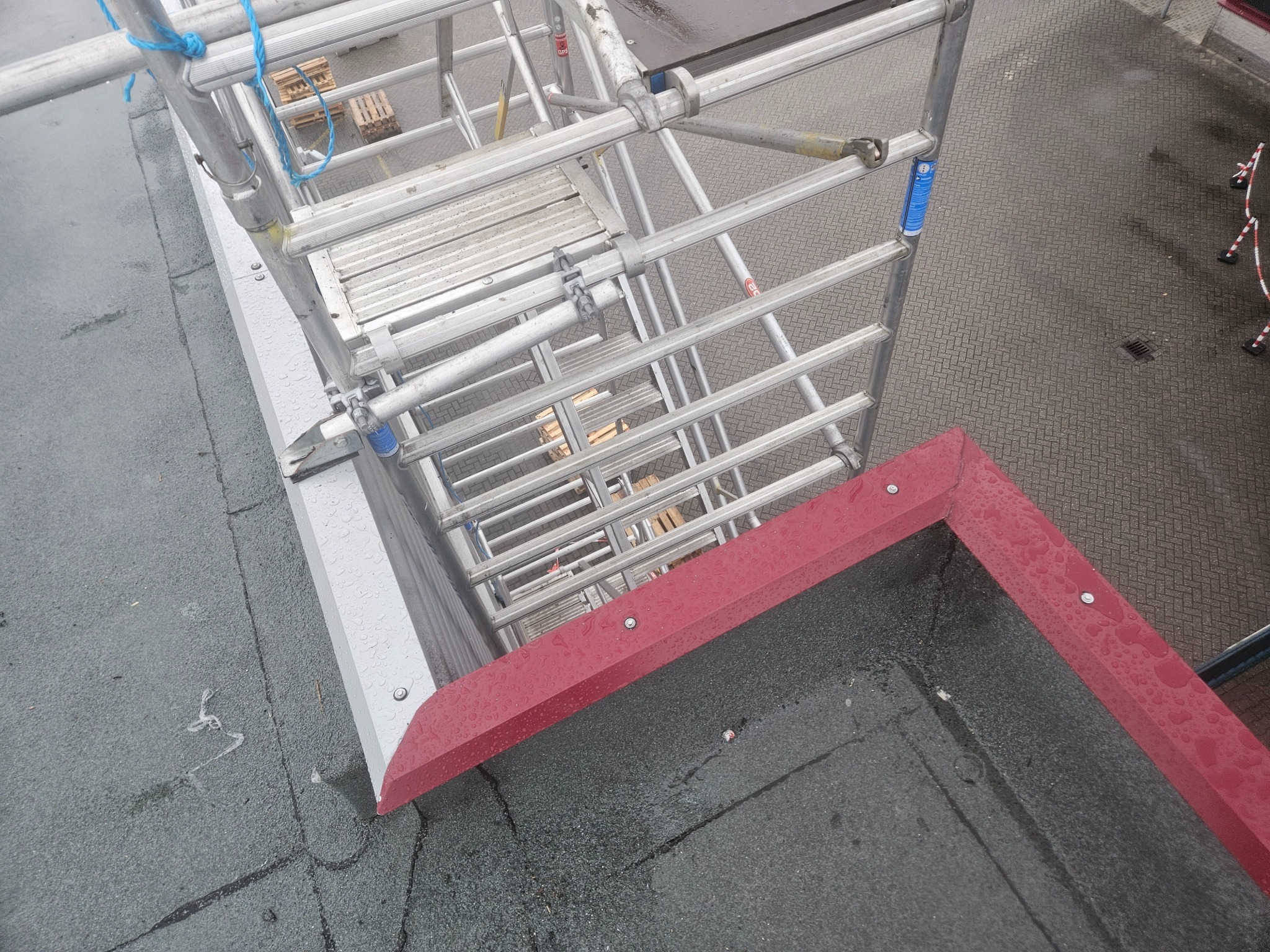 Widok z góry na fragment dachu z papy, z czerwonym obramowaniem i aluminiowe rusztowanie. Widoczne krople wody na powierzchni.