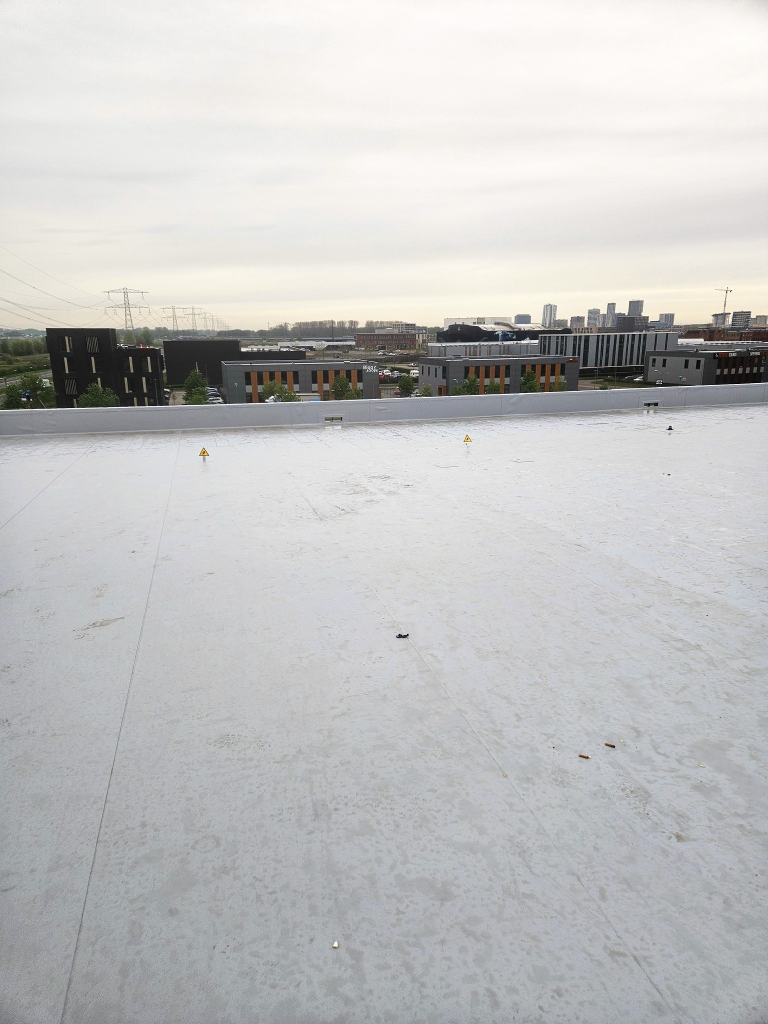 Biała, płaska powierzchnia dachu z widocznymi znakami ostrzegawczymi, budynkami w tle i pochmurnym niebem. Nowe krycie dachu wykonane z membrany PVC.