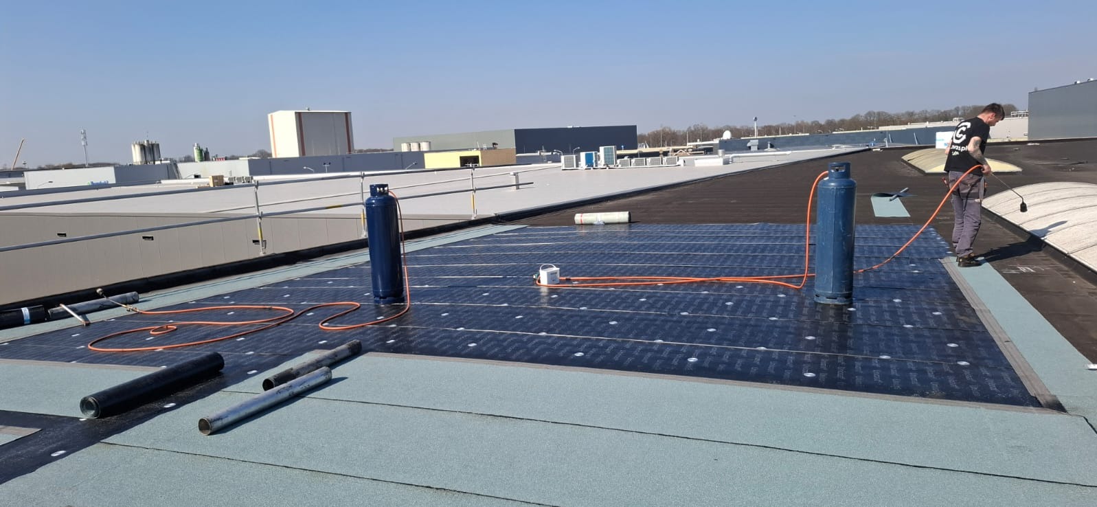 Prace dekarskie na dachu budynku: dekarz z palnikiem, butle gazowe, rolki papy. Widoczne warstwy izolacyjne i materiały przygotowane do montażu na jasnym dachu.