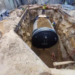 Czarny zbiornik Oksydan w wykopie, instalowany przez dwóch pracowników w kaskach i kamizelkach. Metalowe wzmocnienia i rury w tle. Etap instalacji.