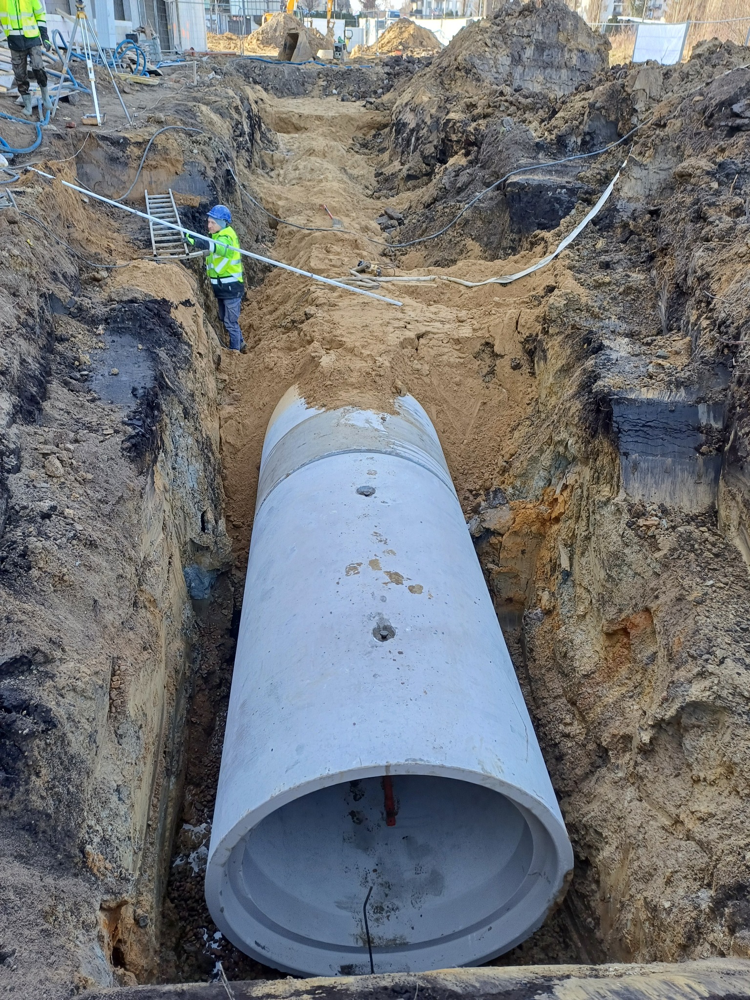 Betonowa rura w wykopie, element instalacji sanitarnej. Pracownik w odblaskowej kamizelce nadzoruje montaż. Widoczne ślady wykopu i ziemia.