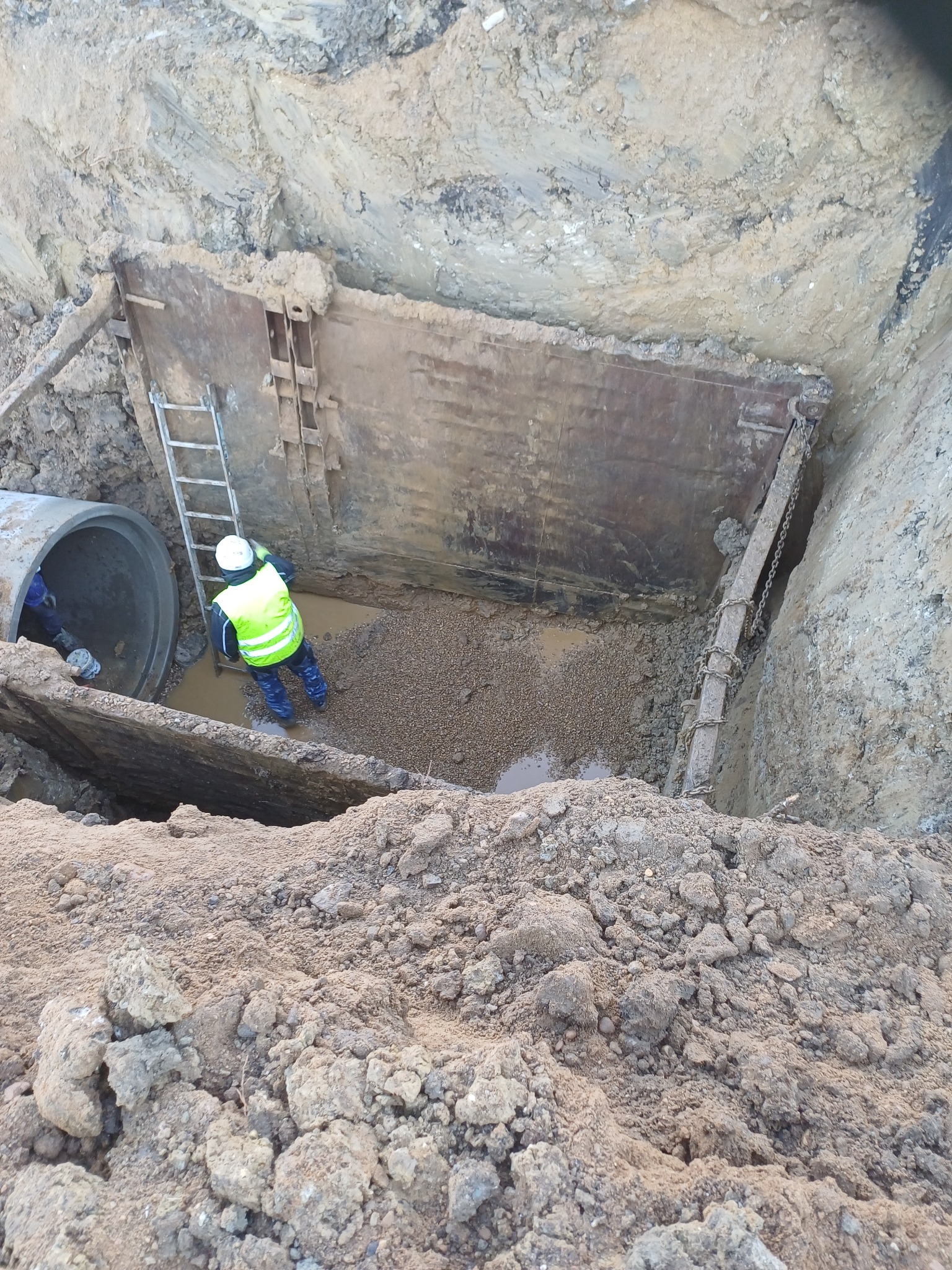 Pracownik w wykopie zabezpieczonym szalunkiem, obok rura kanalizacyjna i drabina. Prace ziemne przy instalacji wod-kan.