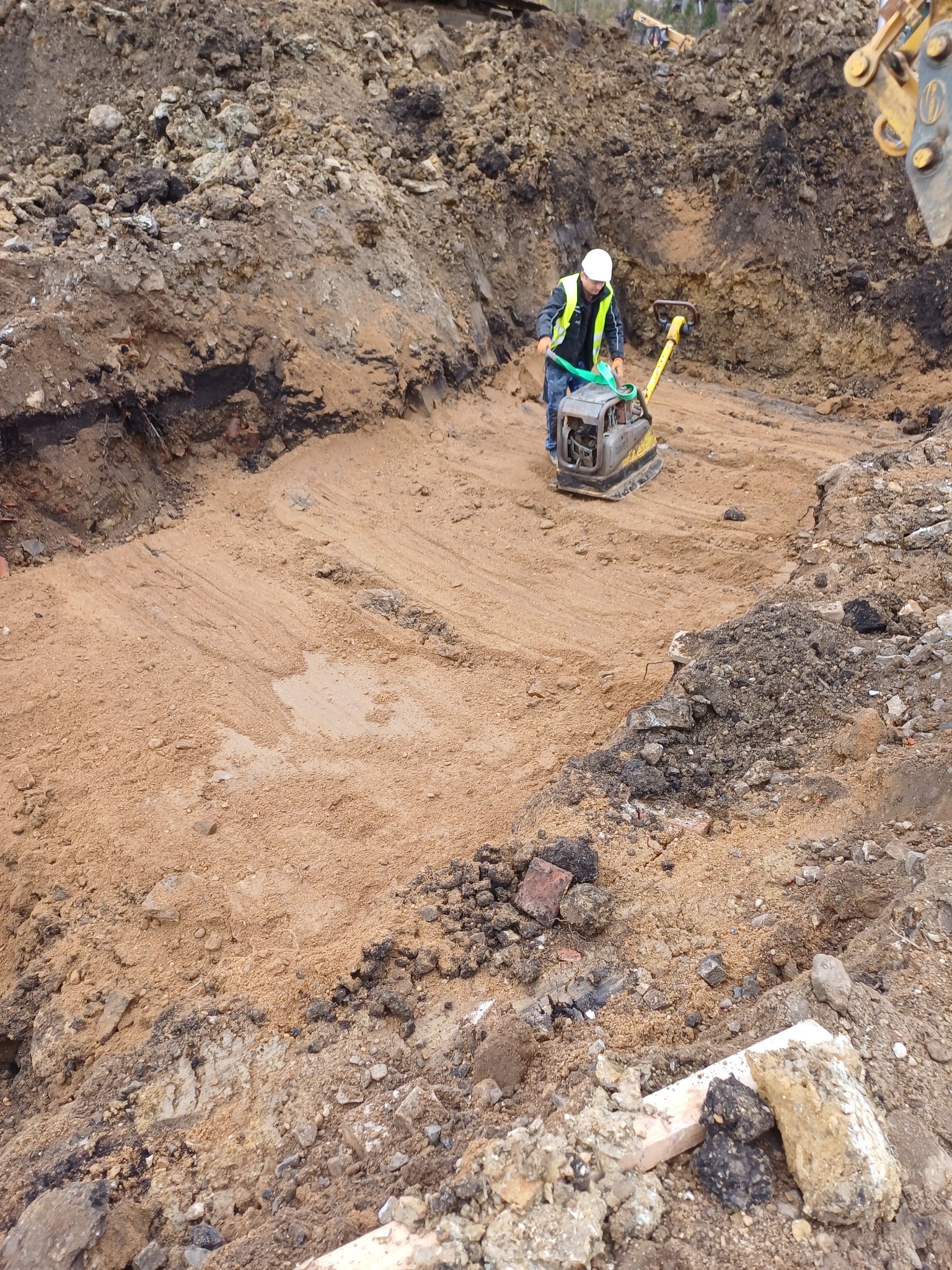 Pracownik w wykopie zagęszcza piasek zagęszczarką wibracyjną. Widoczne ślady pracy maszyny, biały kask i żółta kamizelka odblaskowa.