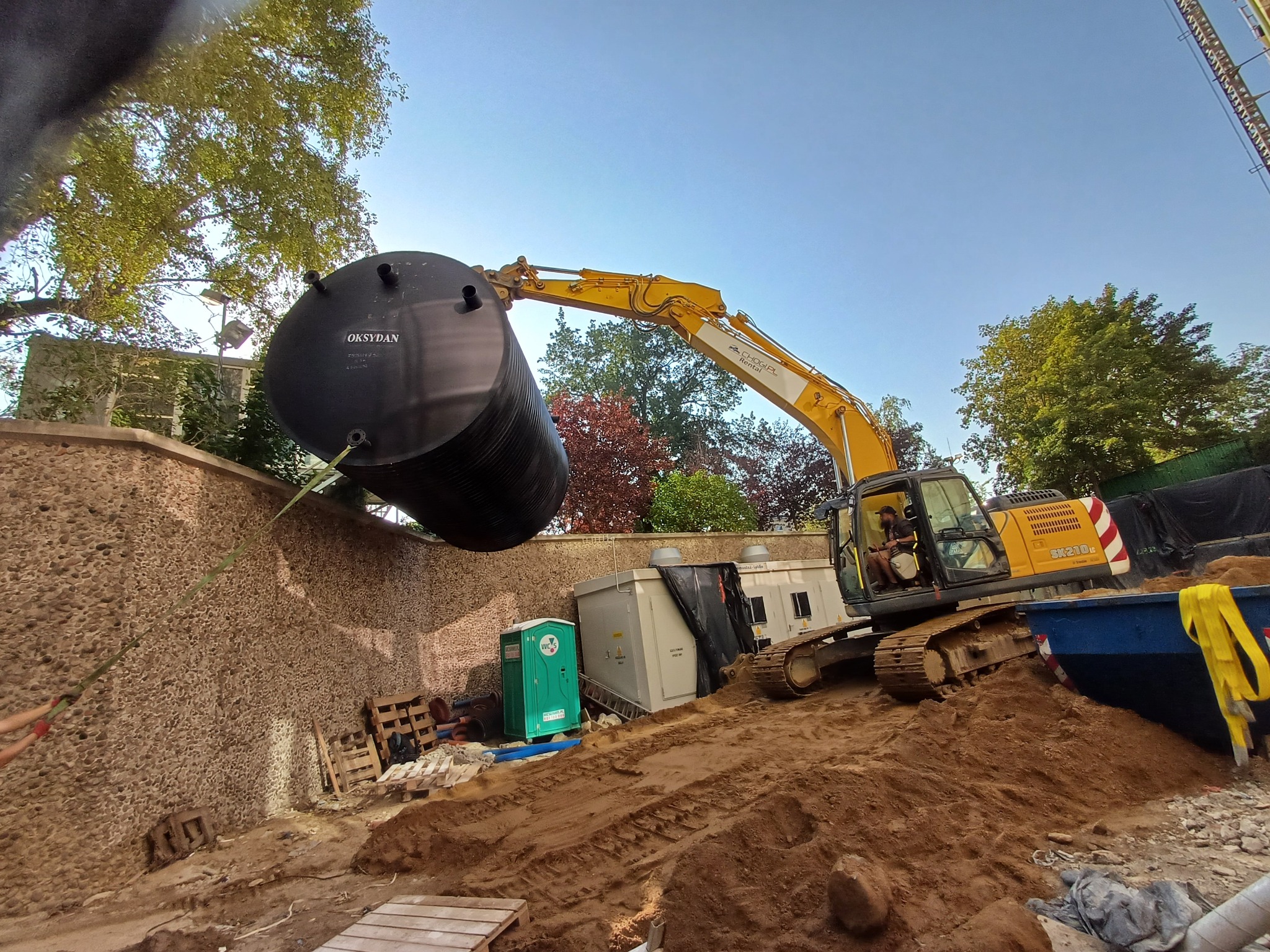 Żółta koparka umieszcza czarny zbiornik Oksydan w wykopie na tle ściany z kamienia i zieleni. Widoczne elementy instalacji i toaleta TOI TOI.