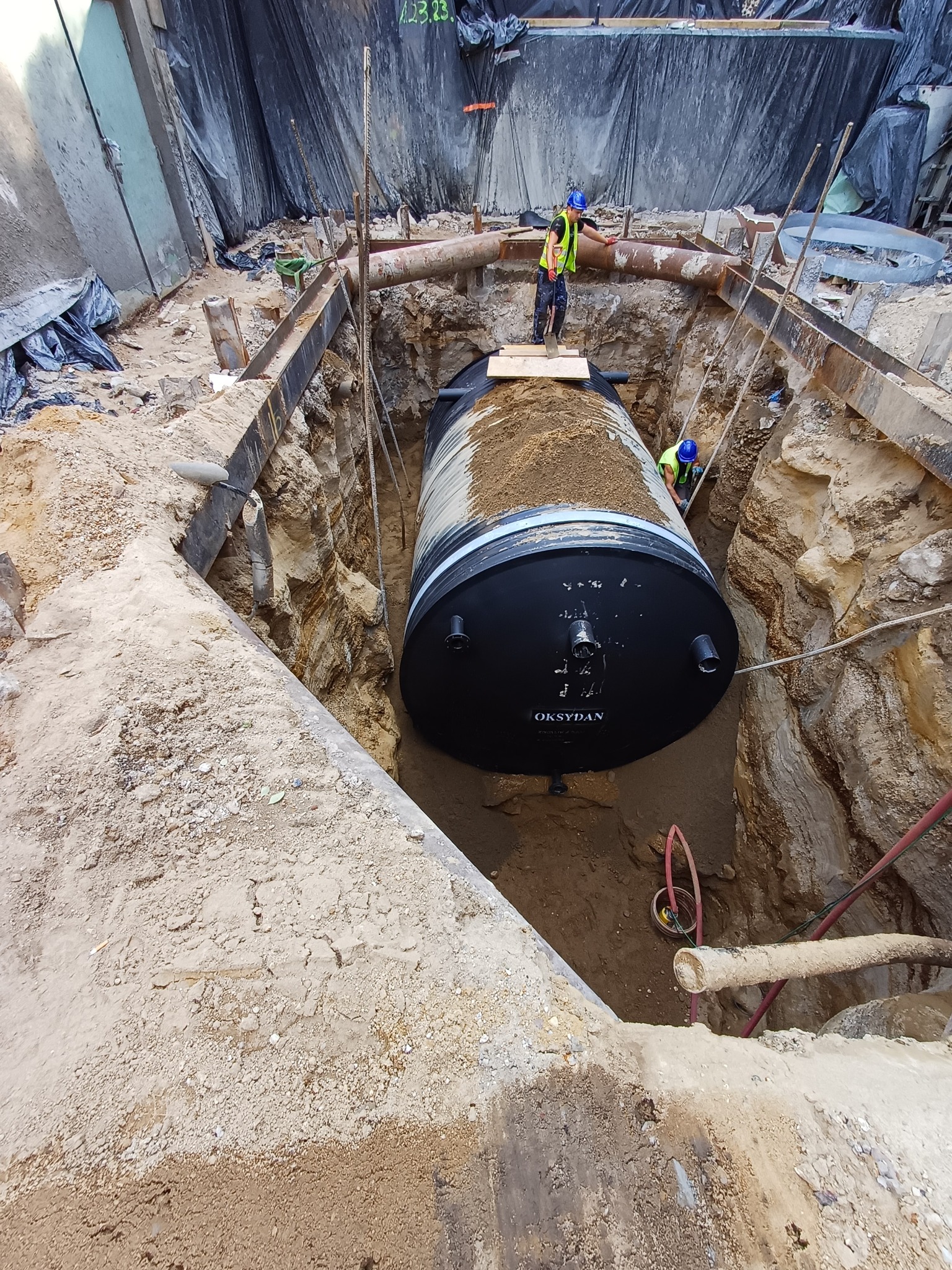 Czarny zbiornik Oksydan w wykopie, instalowany przez dwóch pracowników w kaskach i kamizelkach. Metalowe wzmocnienia i rury w tle. Etap instalacji.