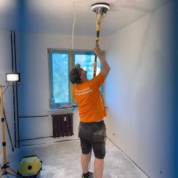 FILBUD - Pracownik w trakcie szlifowania sufitu szlifierką z odkurzaczem w pomieszczeniu po gładziowaniu. Okno zabezpieczone folią. Widoczny sprzęt oświetleniowy i odkurzacz.