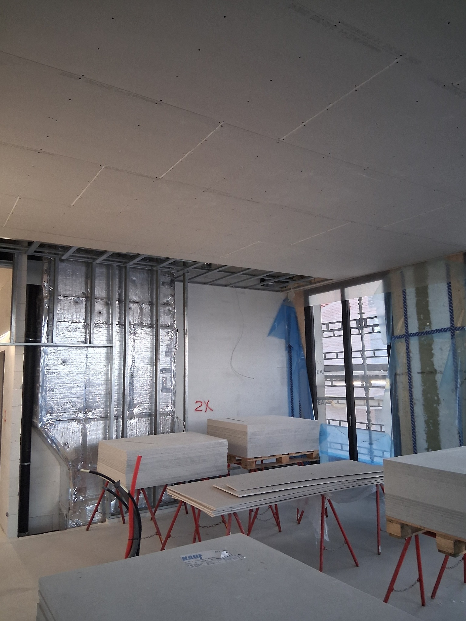 Montaż płyt gipsowo-kartonowych na suficie i ścianach. Widoczne profile metalowe, izolacja i stosy płyt na kozłach. Etap prac wykończeniowych w jasnym wnętrzu.