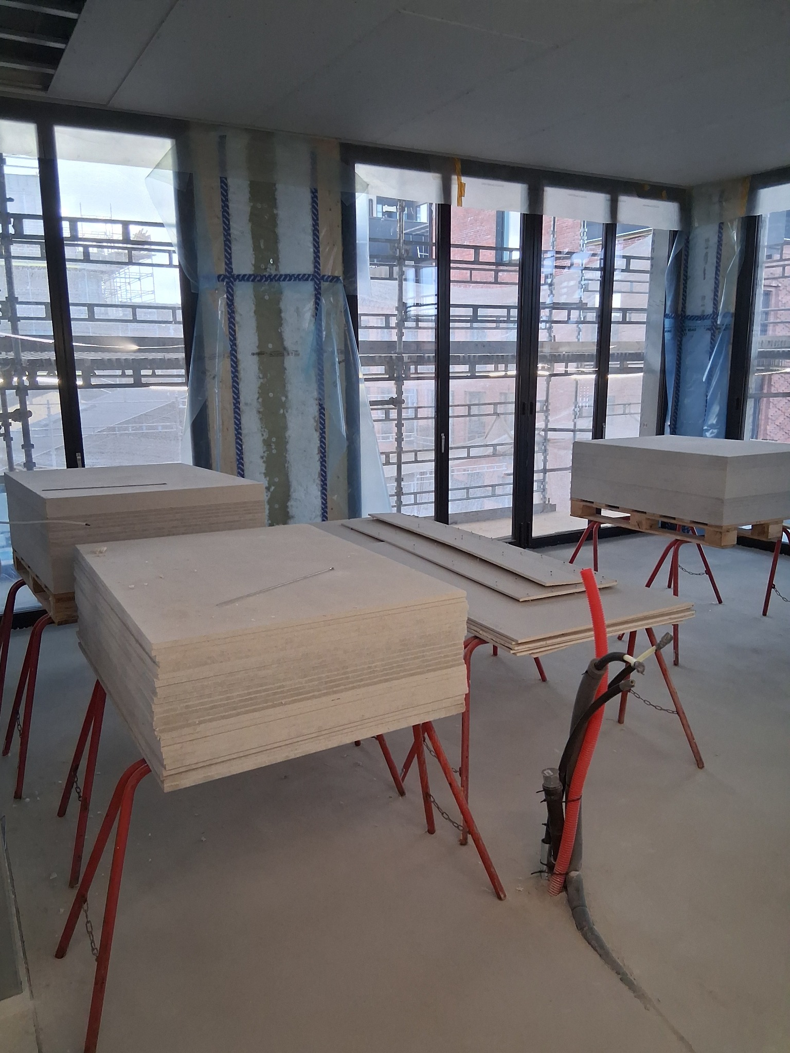 Stosy płyt kartonowo-gipsowych na kozłach w surowym wnętrzu z dużymi oknami. Widoczne instalacje i zabezpieczenia okien folią. Przygotowanie do montażu.