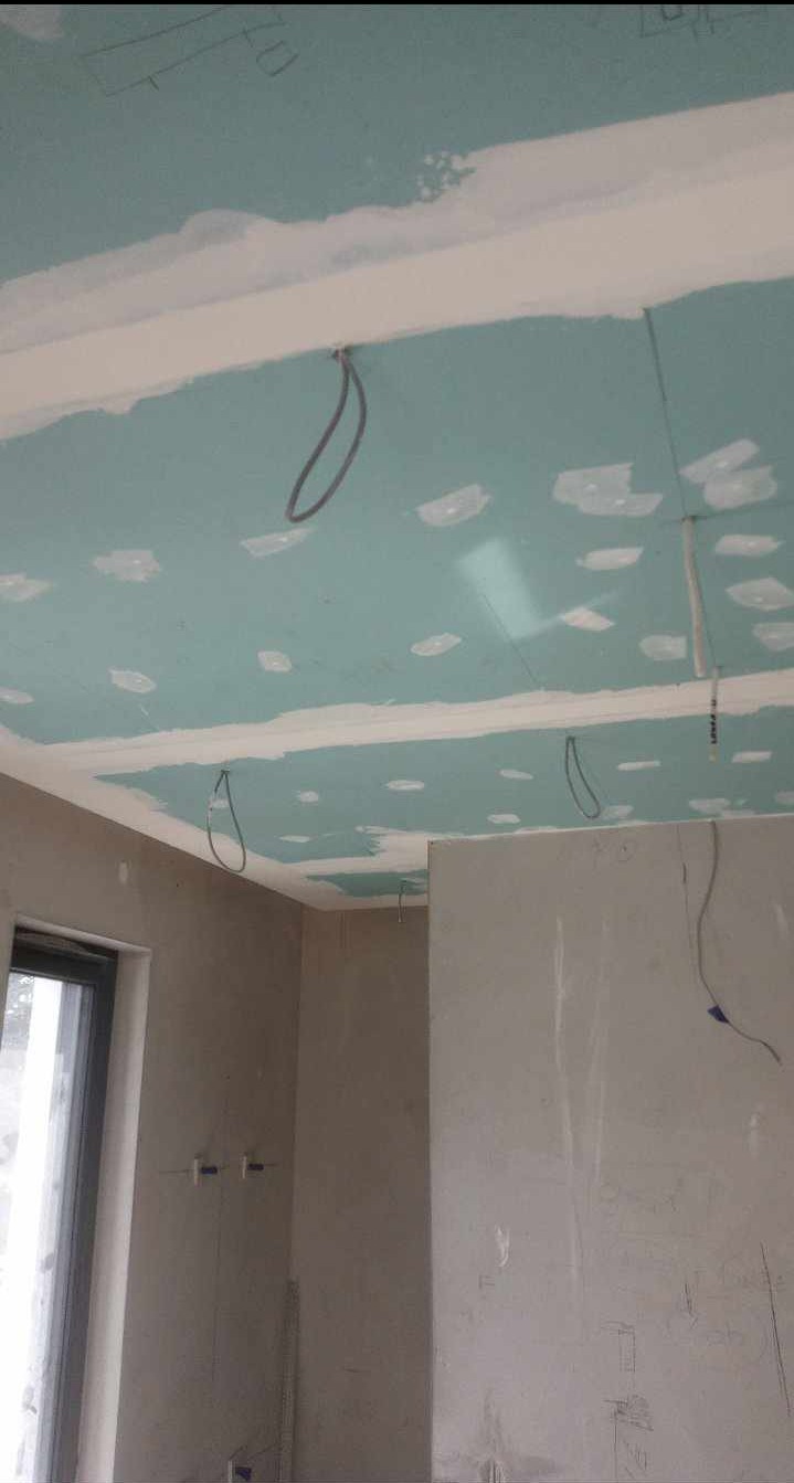 Montaż sufitu z płyty karton-gips w pomieszczeniu z oknem. Widoczne kable wystające z sufitu, przygotowane do instalacji oświetlenia. Szpachlowane łączenia płyt.