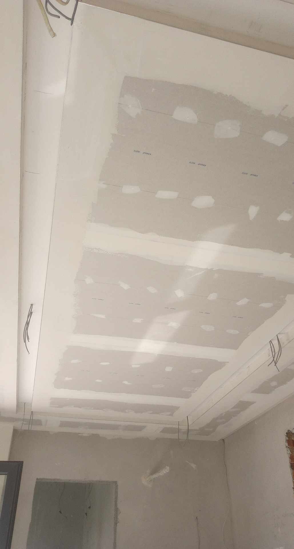 Sufit podwieszany z płyt kartonowo-gipsowych w trakcie szpachlowania, widoczne kable instalacji elektrycznej. Detal wykończenia wnętrza w surowym stanie.