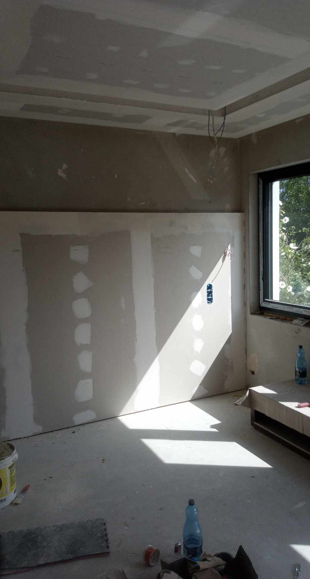 Pokój w trakcie prac wykończeniowych: płyty gipsowo-kartonowe na ścianach, szpachlowany sufit z elementami dekoracyjnymi, okno z widokiem na zieleń.