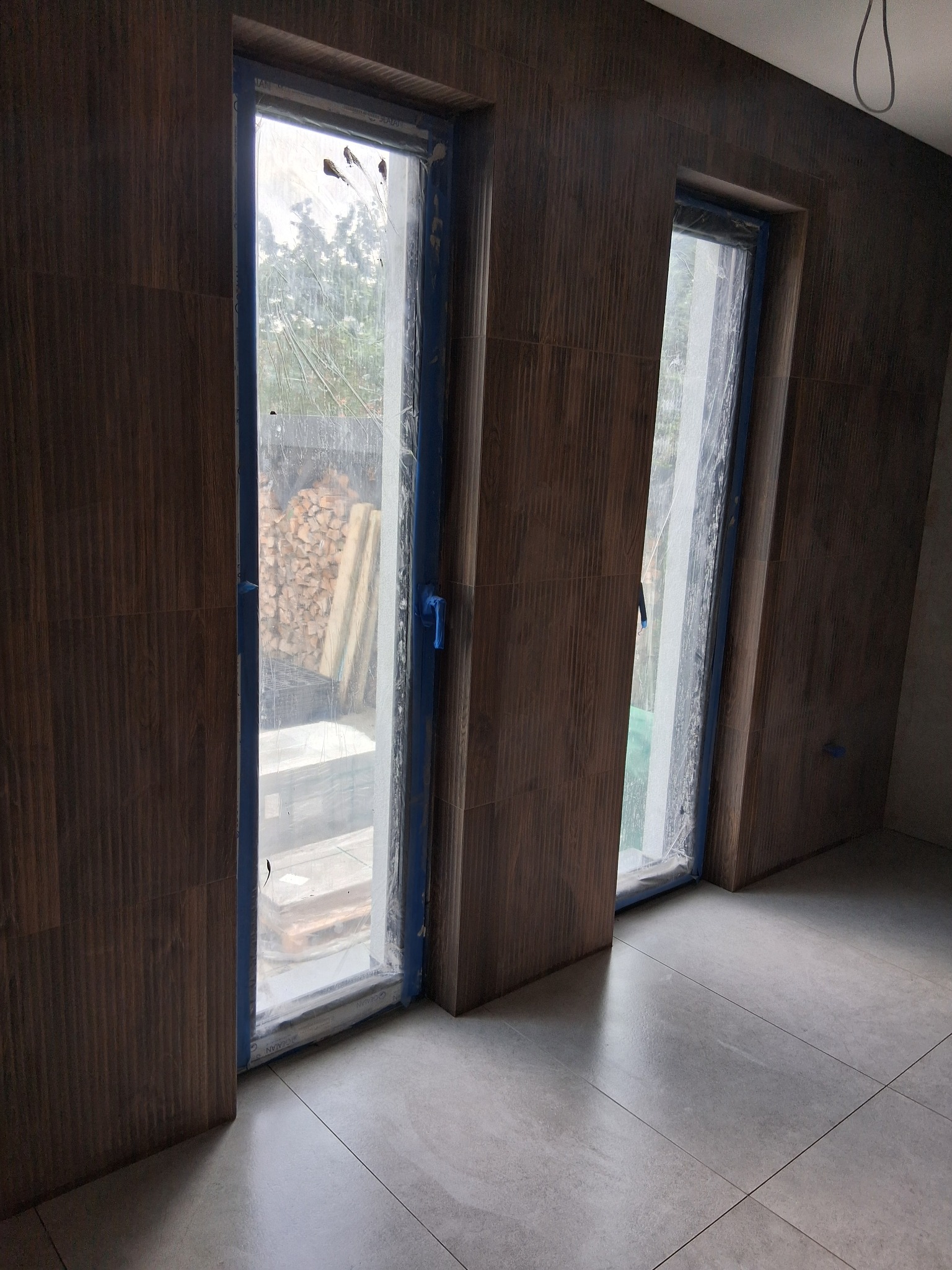 Wykończone wnętrze z drewnopodobnymi płytkami ściennymi i szarą podłogą. Okna zabezpieczone folią malarską. Widoczne detale wykończenia.