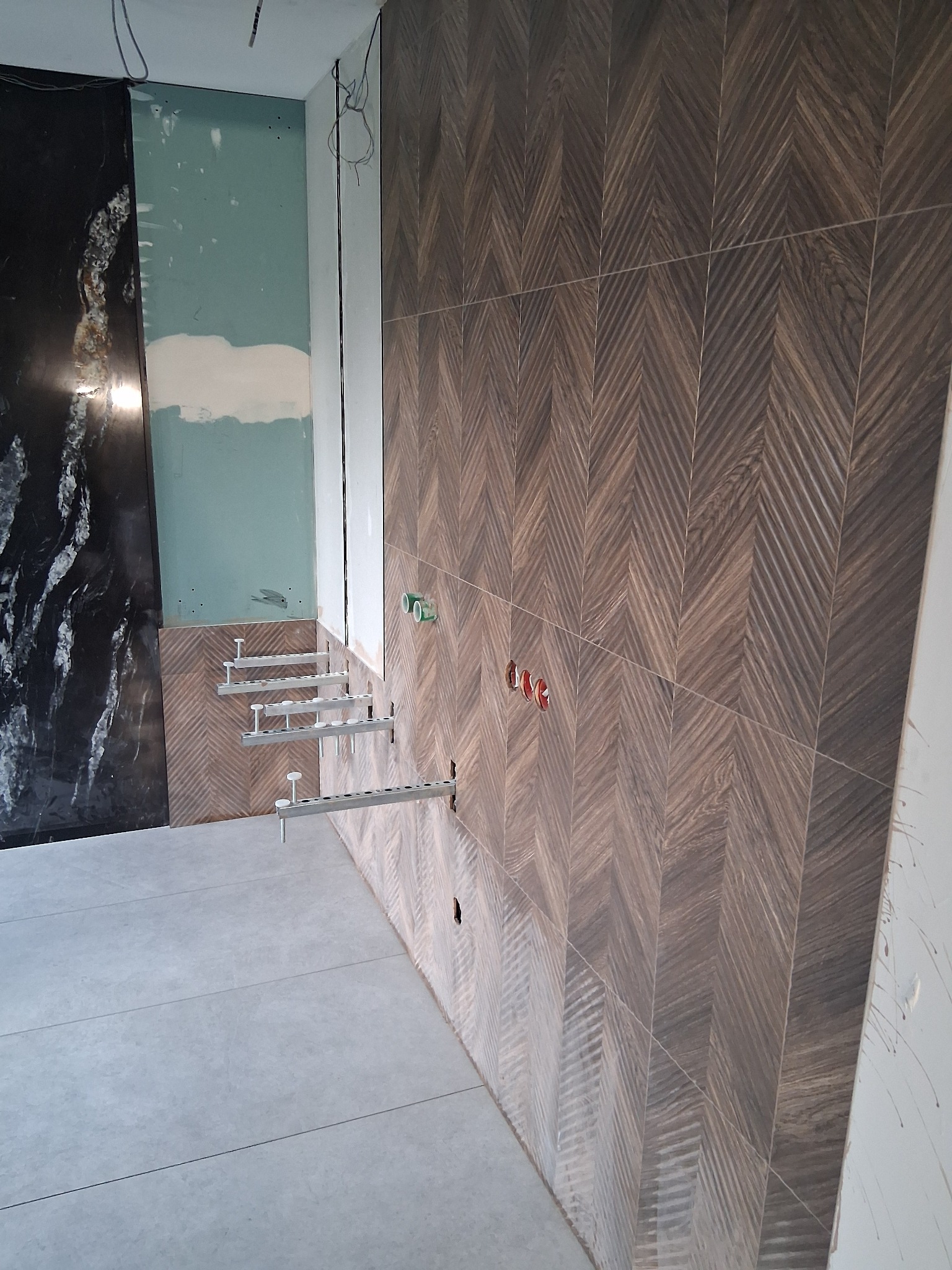 Układanie płytek drewnopodobnych w jodełkę na ścianie, widoczne gniazdka elektryczne i fragment instalacji. Częściowo wykończona ściana z czarnego marmuru.