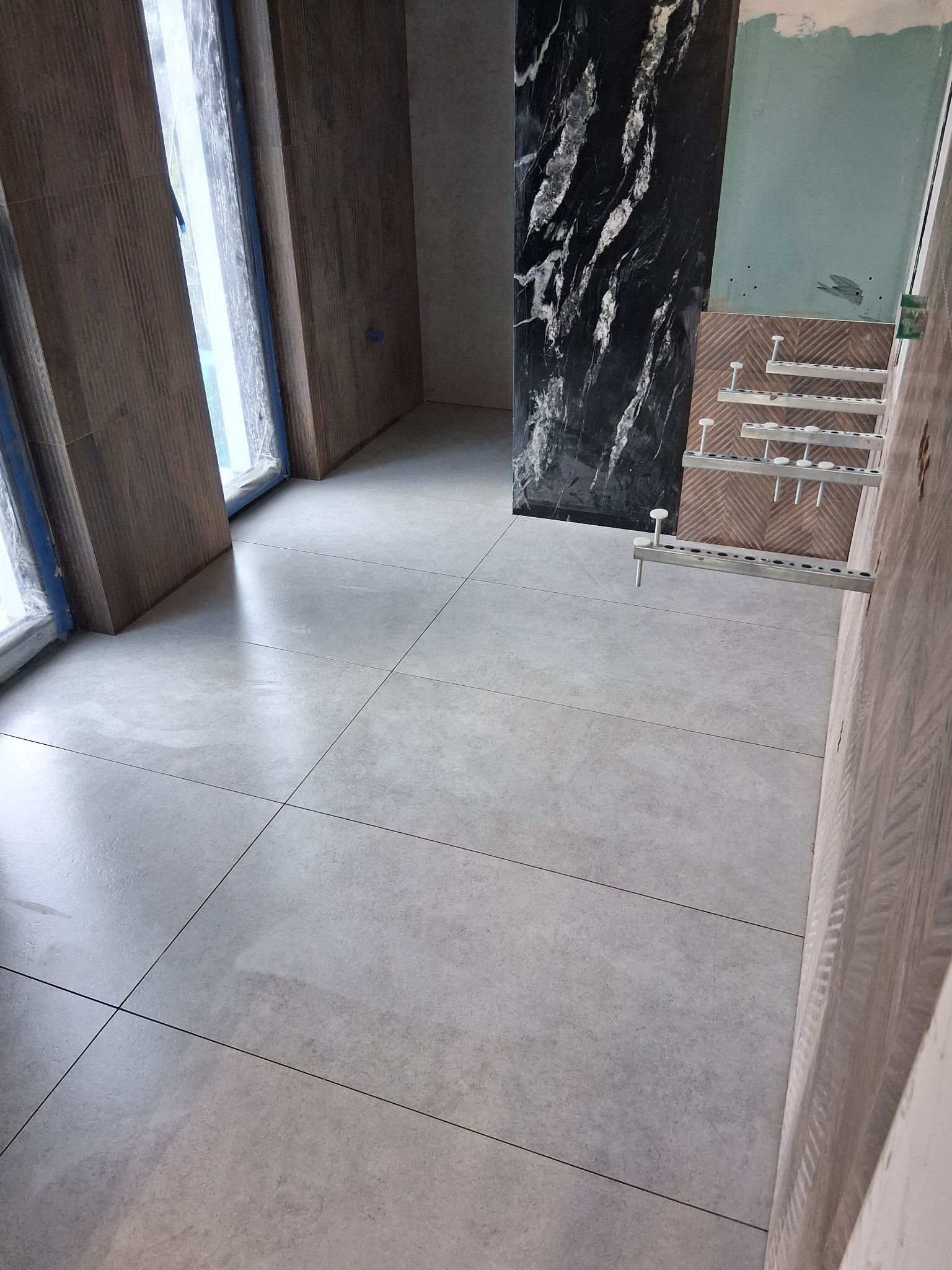 Ułożona szara podłoga z płytek w trakcie prac wykończeniowych. Widoczne fragmenty okna z zabezpieczeniem, ściana z czarnym marmurem i elementy montażowe na ścianie.