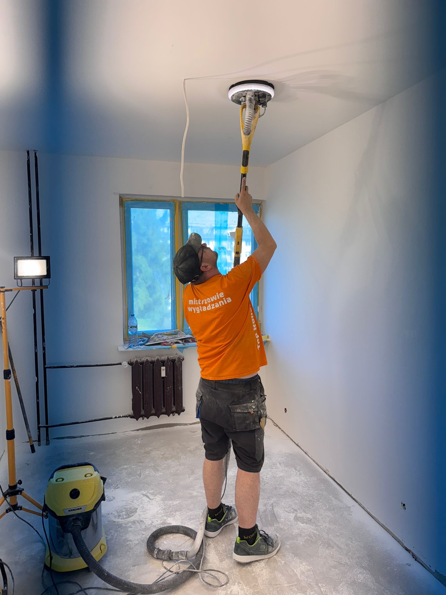 Pracownik w trakcie szlifowania sufitu szlifierką z odkurzaczem w pomieszczeniu po gładziowaniu. Okno zabezpieczone folią. Widoczny sprzęt oświetleniowy i odkurzacz.