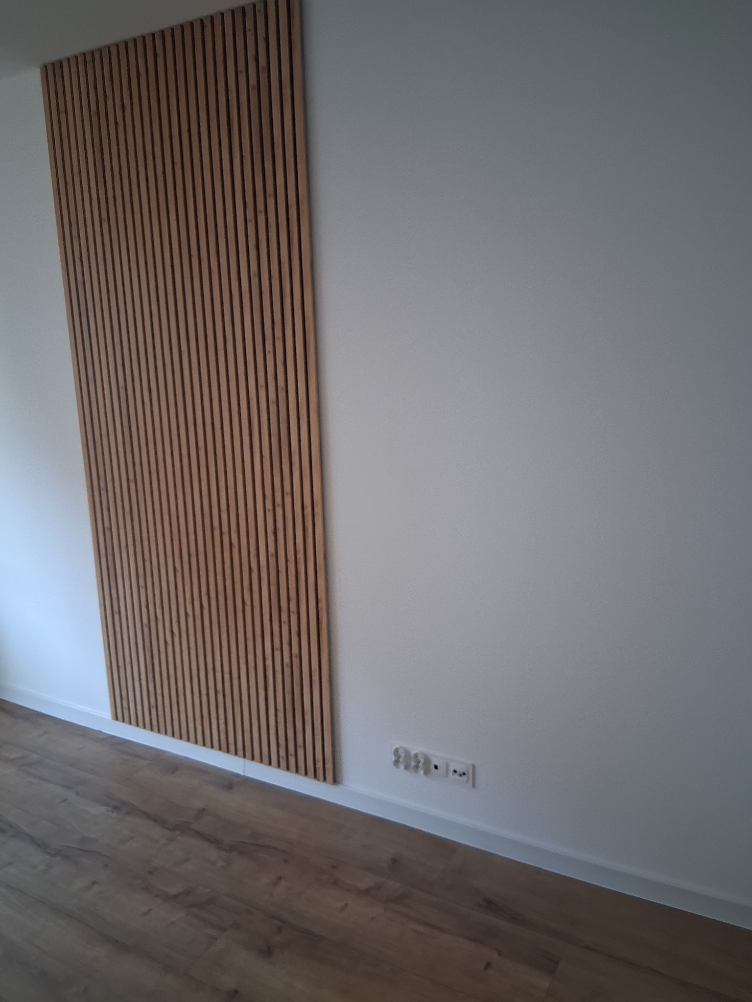 Drewniane lamele dekoracyjne na białej ścianie, widoczne gniazdka elektryczne i fragment podłogi z paneli. Nowoczesny design wnętrza.