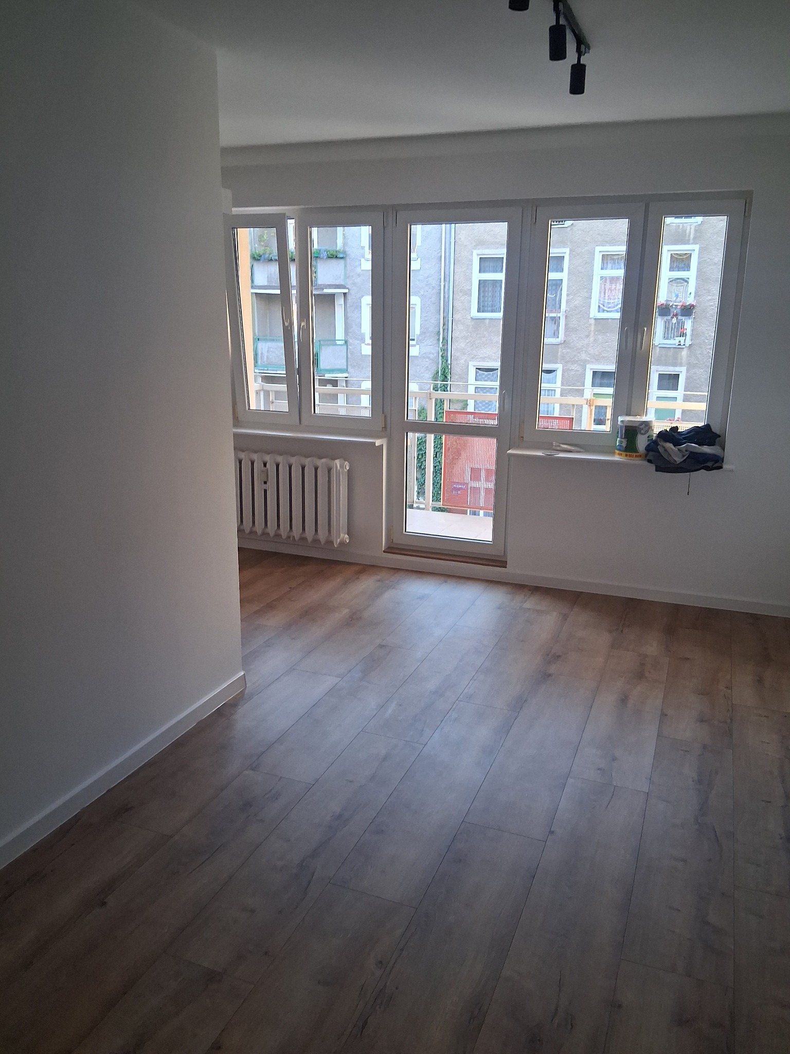 Jasne, odświeżone wnętrze pokoju po remoncie. Nowa podłoga z paneli, świeżo pomalowane ściany na biało, okno z widokiem na budynek mieszkalny. Grzejnik pod oknem.