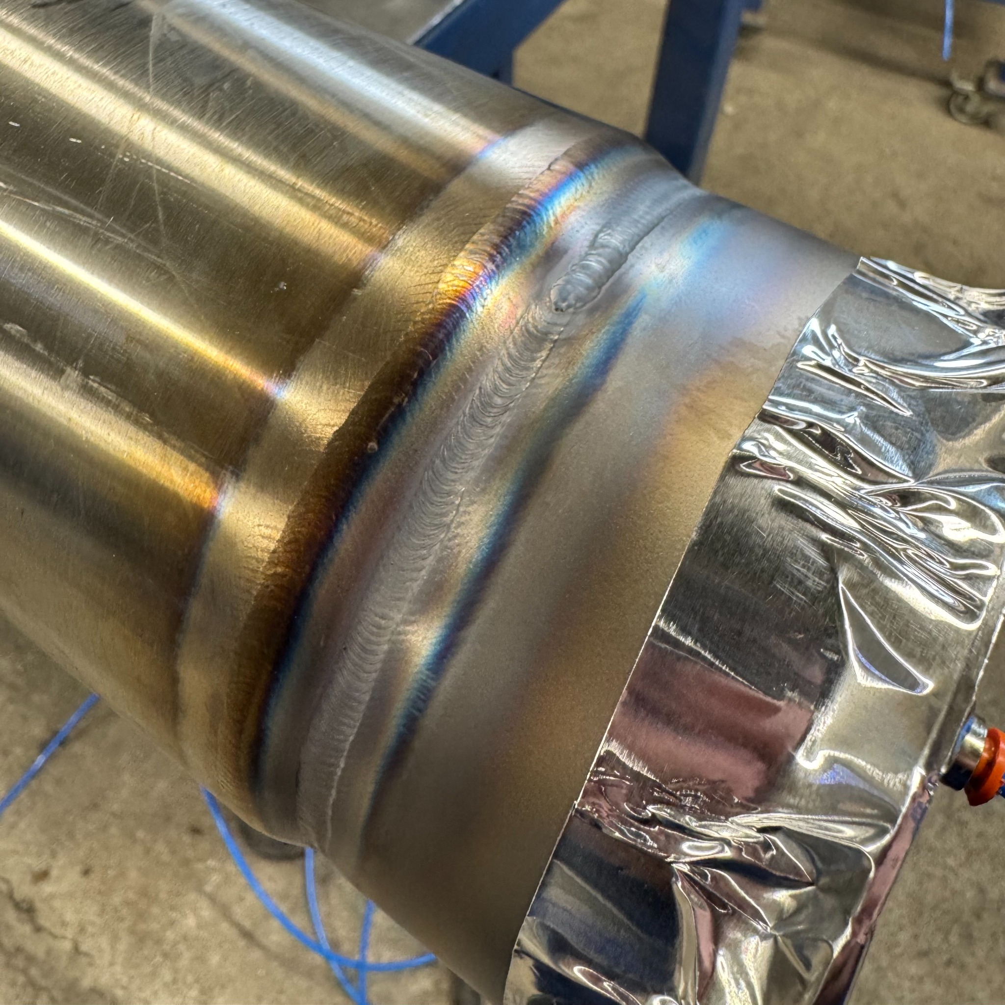 Szlifowany, metalowy cylinder z widocznym spoiwem spawalniczym i tęczowymi przebarwieniami termicznymi. Część cylindra owinięta folią ochronną.